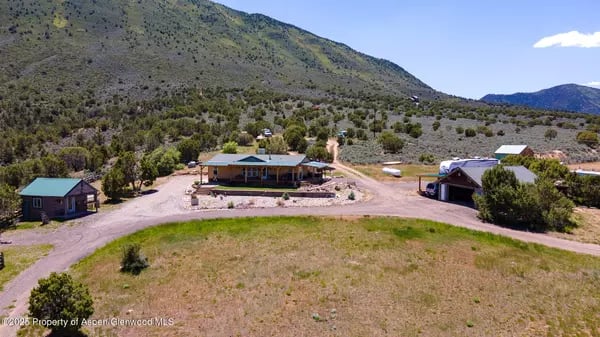 Aerial view of a rural home and outbuildings below a scrub-covered mountain.