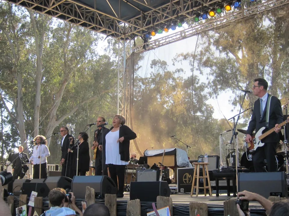 Mavis Staples in white shirt and black cardigan performing center stage with gospel choir and band, backed by large projection screen showing trees, with colorful stage lights overhead