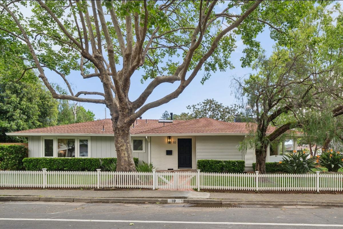 Single-story house behind a white picket fence, shaded by large mature trees.
