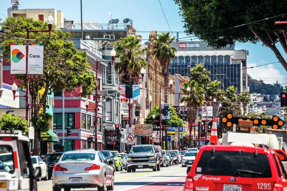 The Mission District in San Francisco, featuring a bustling street lined with colorful buildings, palm trees, and a mix of cars and pedestrians. A US Bank building is visible in the background, along with vibrant storefronts and street banners.