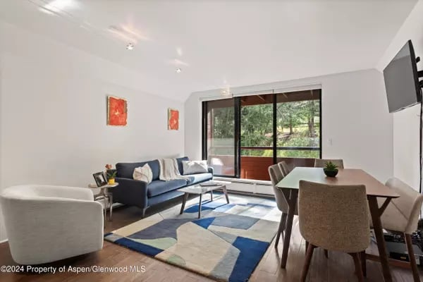 Living and dining room with a blue sofa, geometric rug, and glass doors overlooking trees.