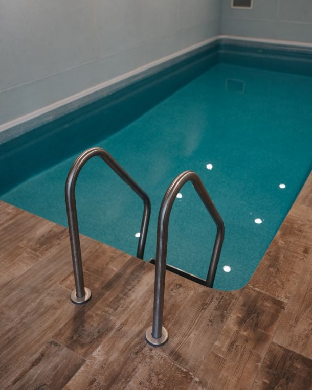 Indoor swimming pool with teal water, a stainless-steel ladder, and wood-look tile decking.