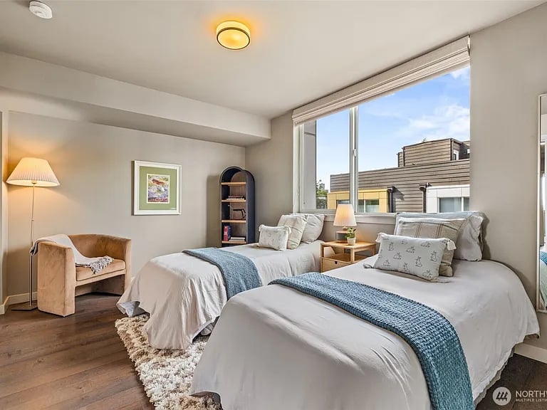 Staged bedroom with two white beds, blue throws, and large windows overlooking a nearby building.