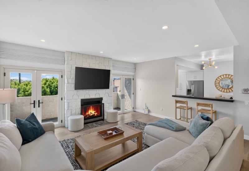 Living room with a stone fireplace, wall-mounted TV, and French doors opening to a tree-lined balcony.