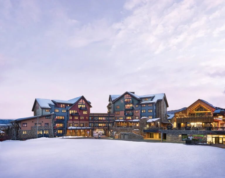 Alpine resort buildings at dusk, glowing windows set against snow-covered roofs and a snowy foreground.