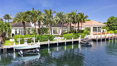 Waterfront home with palm trees, a long dock, boat lifts, and boats on a canal.
