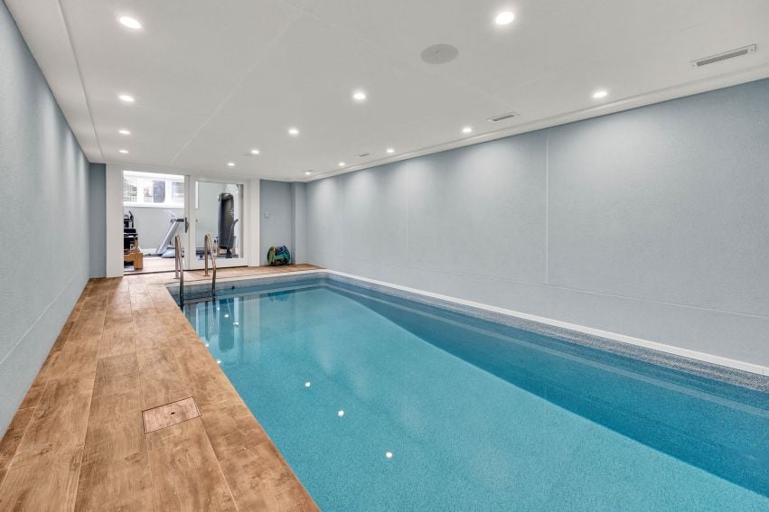 Indoor swimming pool with a wood-look tile deck and exercise equipment beyond glass doors.