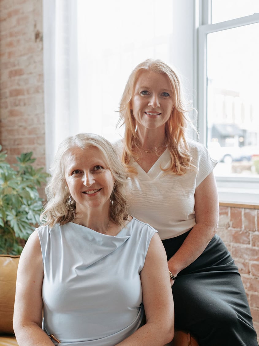 Two smiling women pose together indoors, one seated in a pale blue top and the other standing behind her.