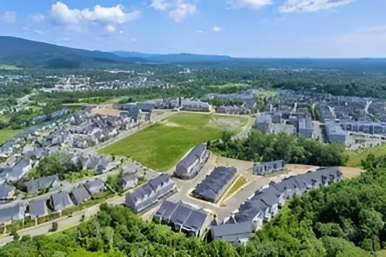 Aerial view of a residential neighborhood around a broad green space, with wooded mountains beyond.