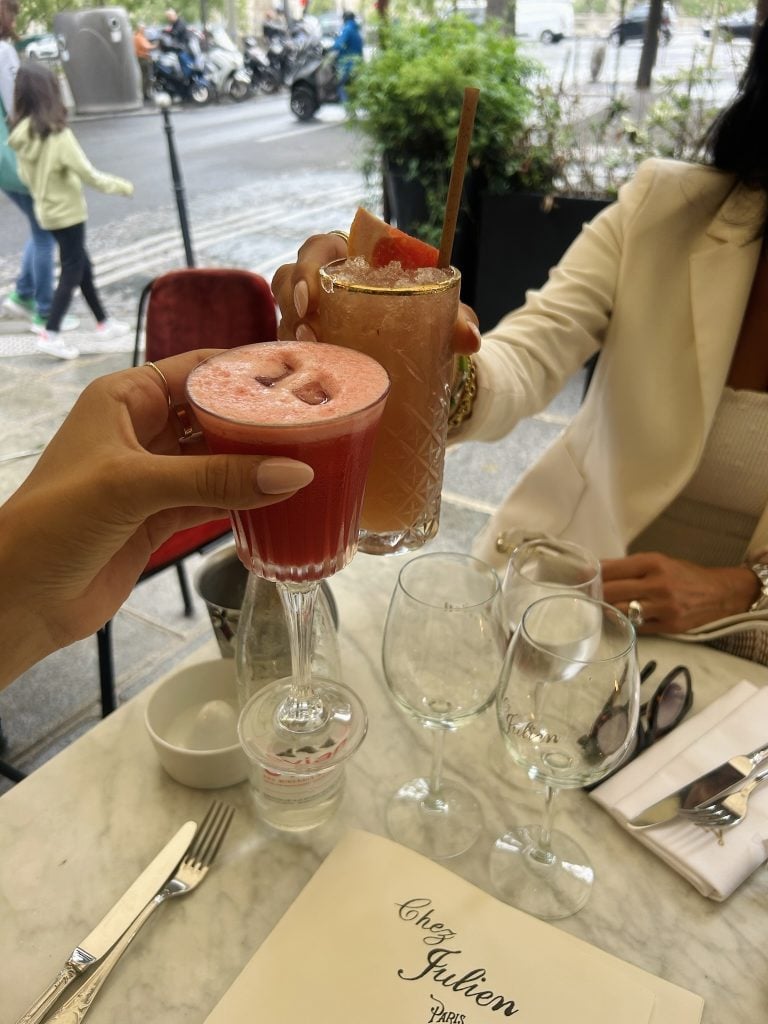 Two cocktails raised over a marble café table beside a Paris sidewalk.