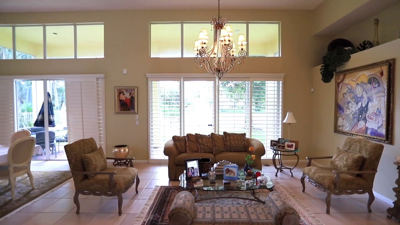 Furnished living room with a tall ceiling, ornate chandelier, and large shuttered windows.