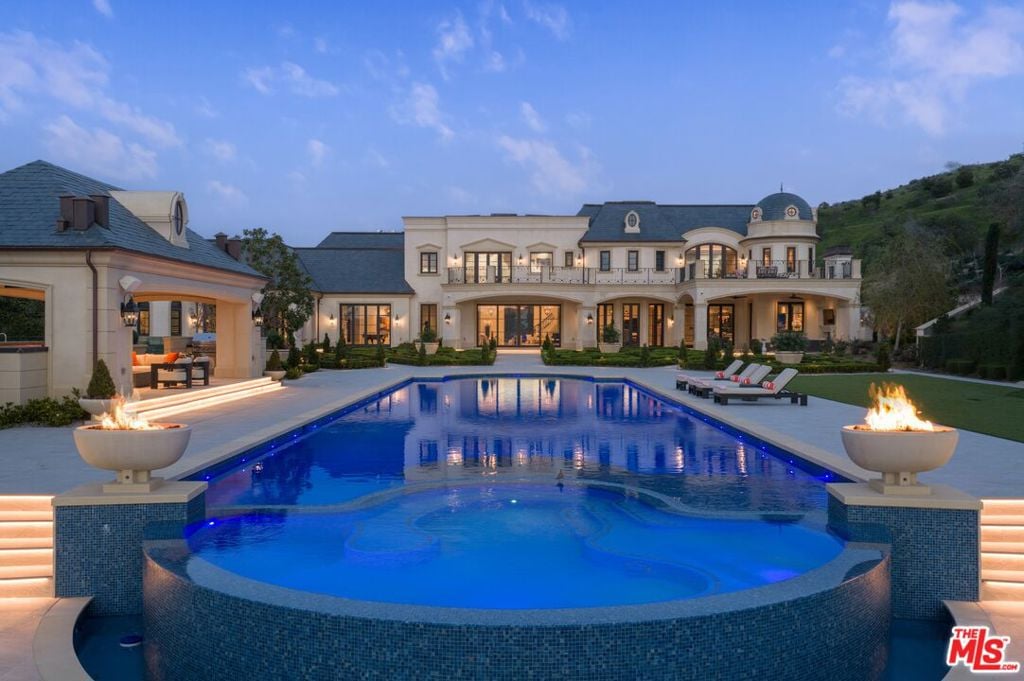 Estate swimming pool at dusk with a raised spa, glowing fire bowls, and a large villa behind it.