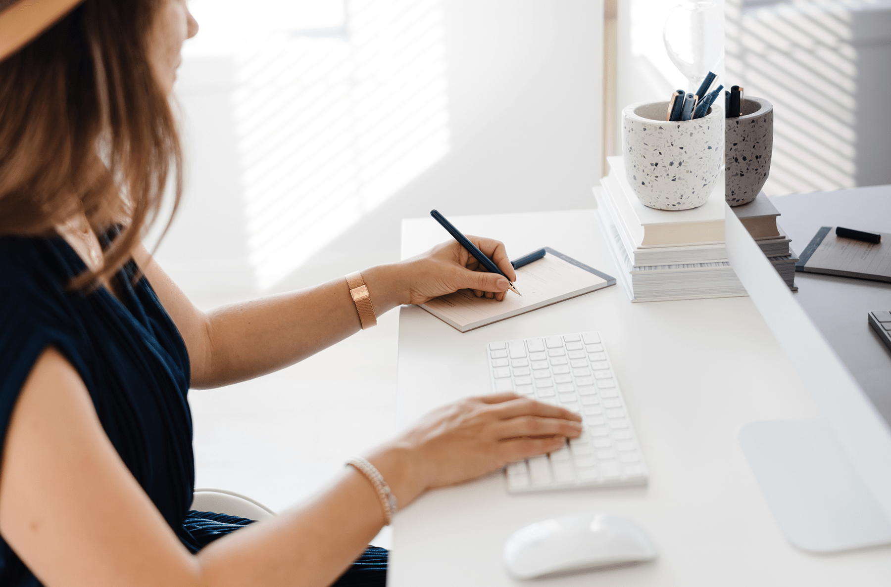 Woman writing in a notebook at a bright desk beside a keyboard and pen holders.
