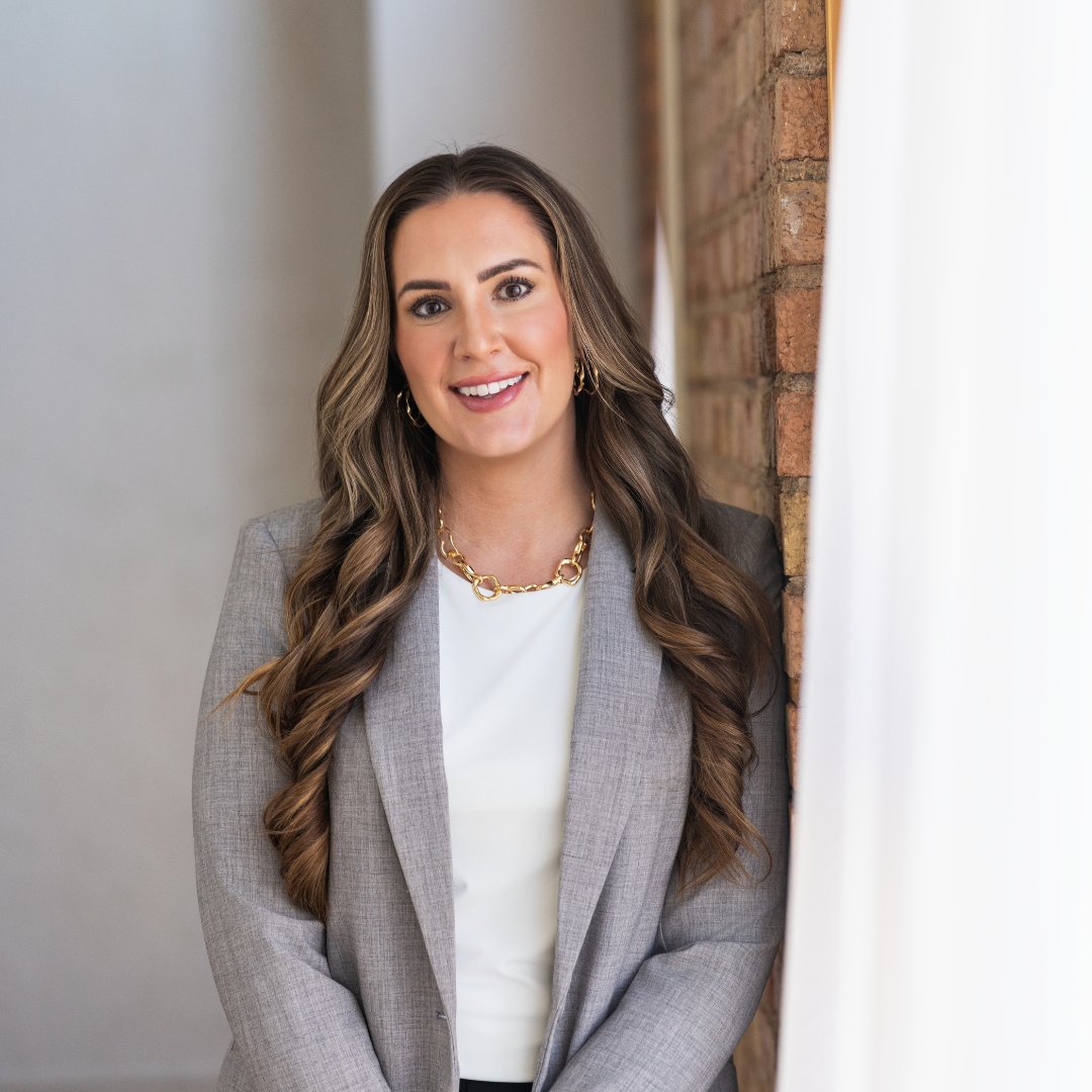 Asha Vonburg, smiling in a gray blazer beside an exposed brick wall.