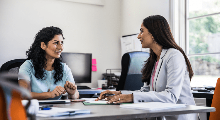 Two women sitting at a desk in an office, smiling and engaged in a conversation. Papers and a computer are visible, conveying a collaborative atmosphere.