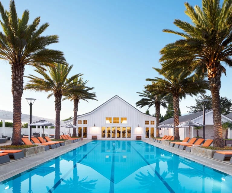Club pool lined with palm trees and orange lounge chairs, facing a white gabled clubhouse.
