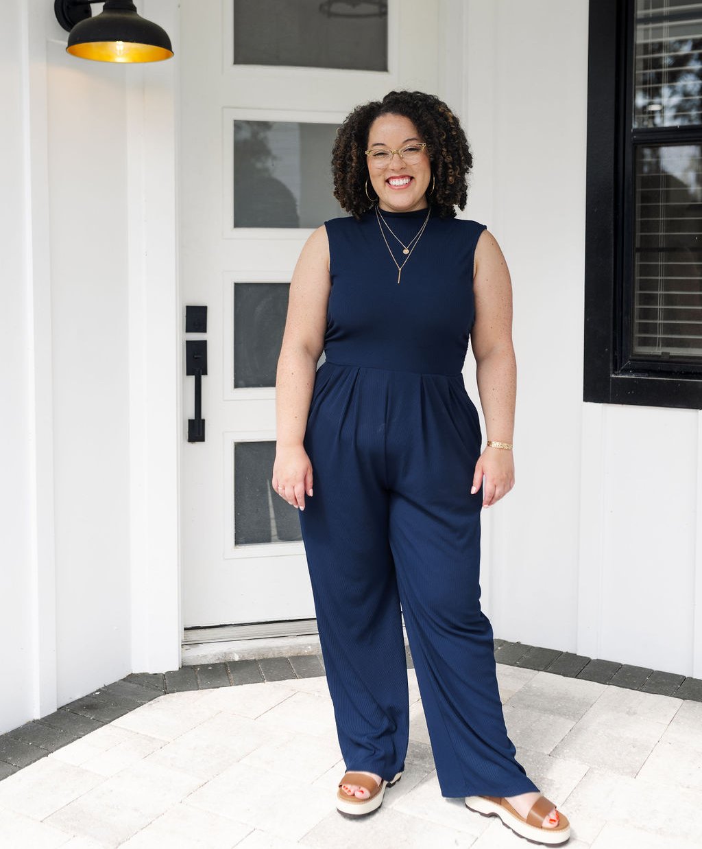 Woman in a navy jumpsuit and tan sandals standing on a porch in front of a white door.