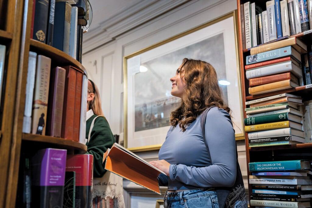 A woman browses a bookstore shelf while holding an open book.
