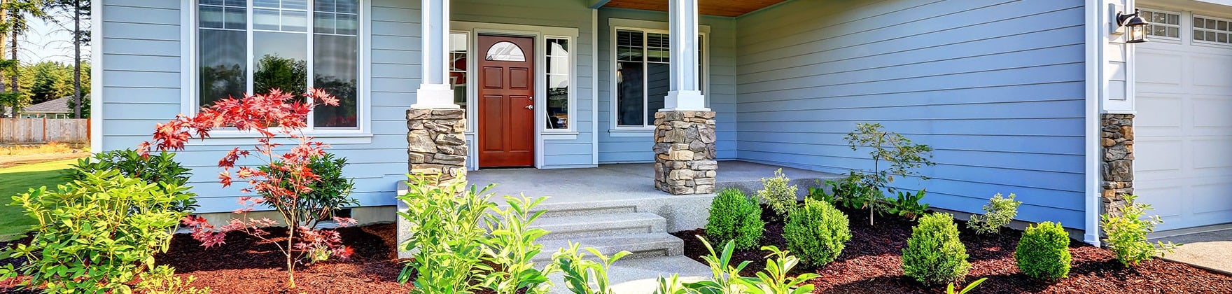 Blue-sided house with a red front door, stone-based porch columns, and landscaped shrubs.