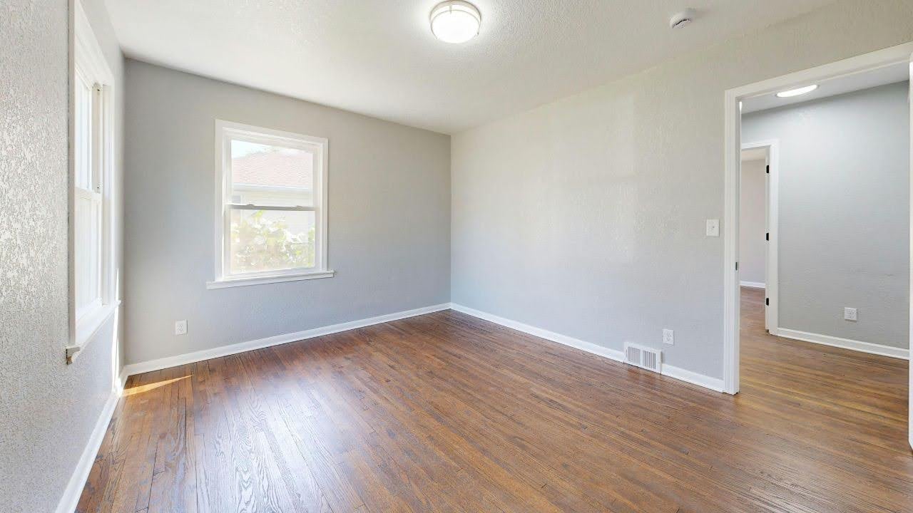 A bright, empty room with light gray walls, wooden floors, and a single window. An open doorway leads to another room.