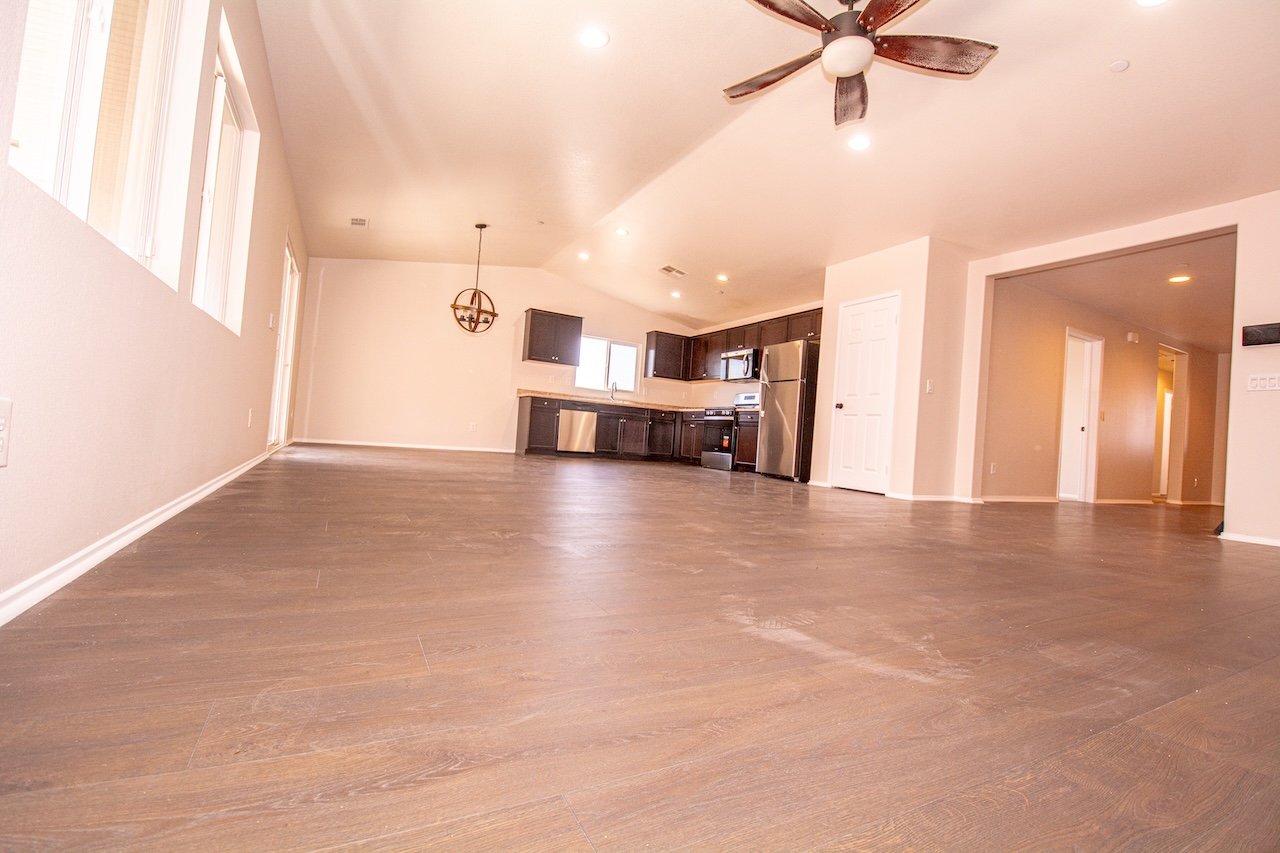Empty open-plan living room and kitchen with wood-look tile floors, dark cabinets, and stainless steel appliances.