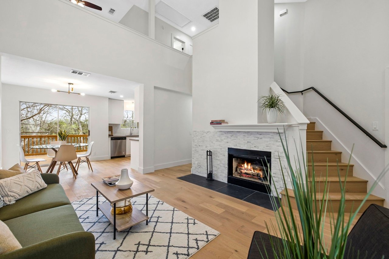 Living room with a stone fireplace, open staircase, and dining area beside large windows.