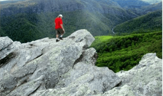 Hiker in a red shirt on a rocky overlook above forested mountain ridges.