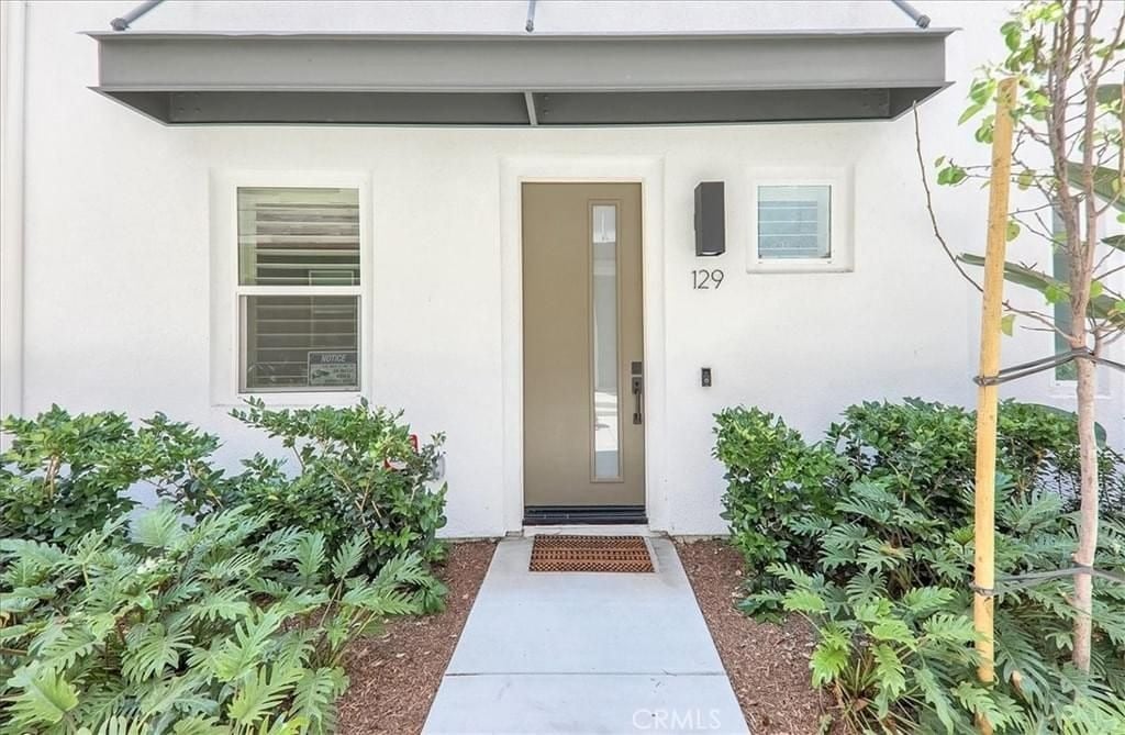  A clean, modern home entrance with a light brown front door, a small walkway, and green bushes on either side.
