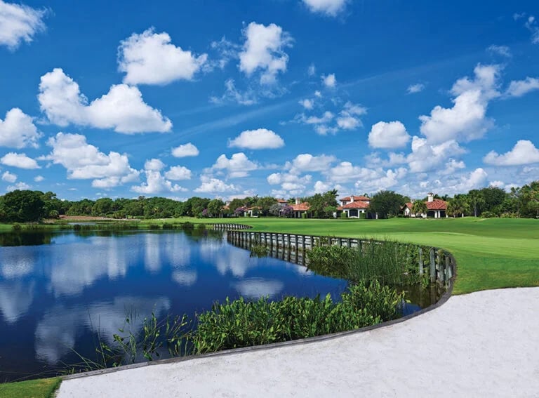 Golf course beside a calm lake, with reeds and homes in the distance.