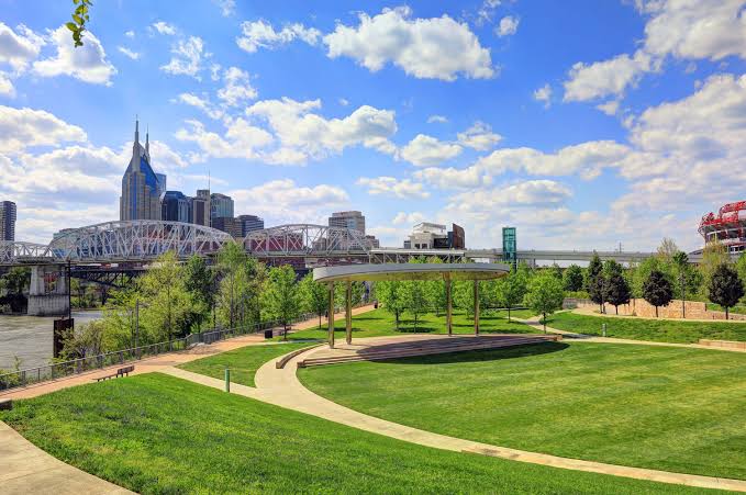 Nashville skyline beyond a riverfront park with green lawns, curving paths, a pavilion, and a bridge.