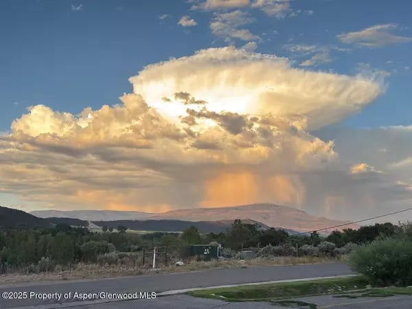 Foothills beneath towering sunlit storm clouds, viewed across a valley from beside a road.