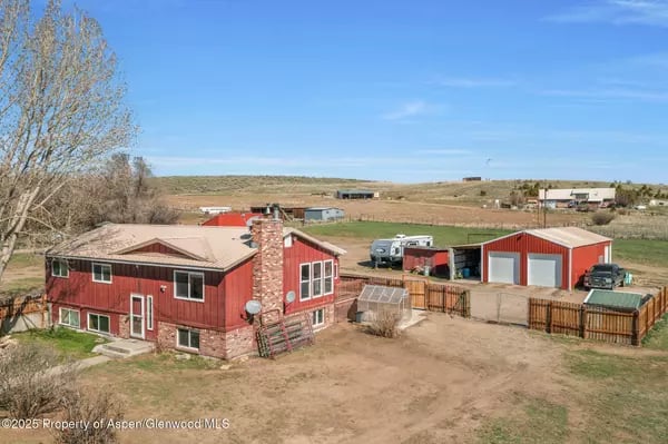 Aerial view of a red rural home with a stone chimney, fenced yard, and detached outbuildings.