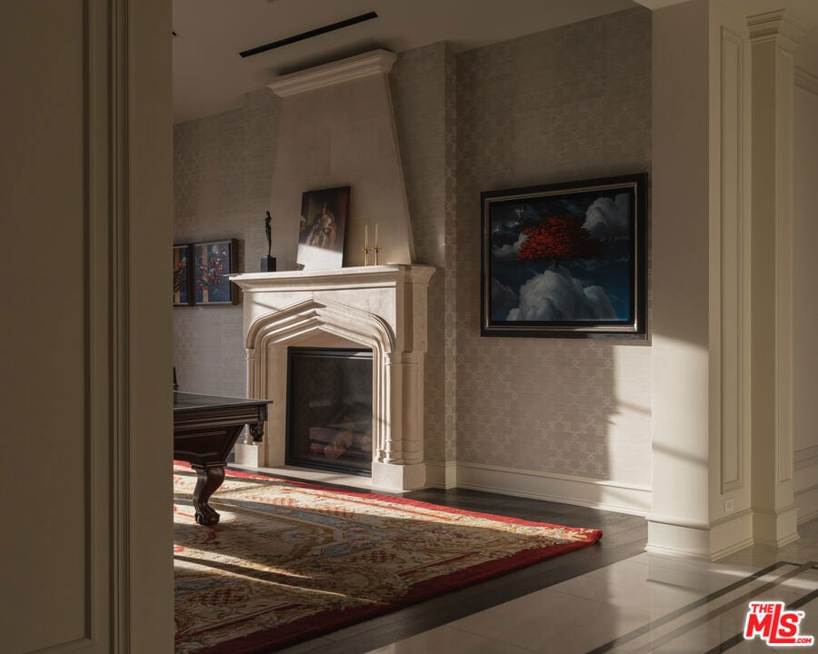 Living room with a sculpted stone fireplace, patterned wallpaper, and sunlight across a red-bordered rug.