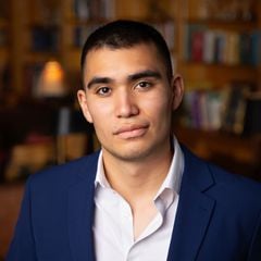 Levi Gonzales in a navy suit and white shirt, with bookshelves softly blurred behind him.