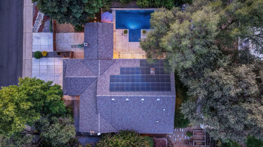 Aerial view of a home with rooftop solar panels, a pool, and a paved patio.
