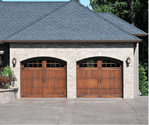 Light-brick garage with two wooden carriage-style doors and a paved driveway.