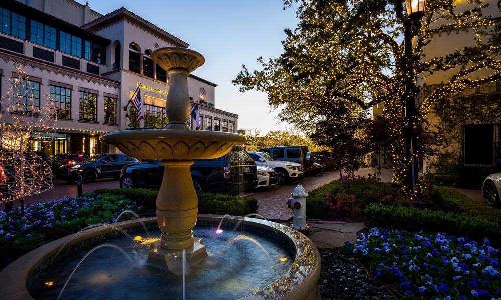 Highland Park Village fountain and flower beds beneath trees strung with lights at dusk.