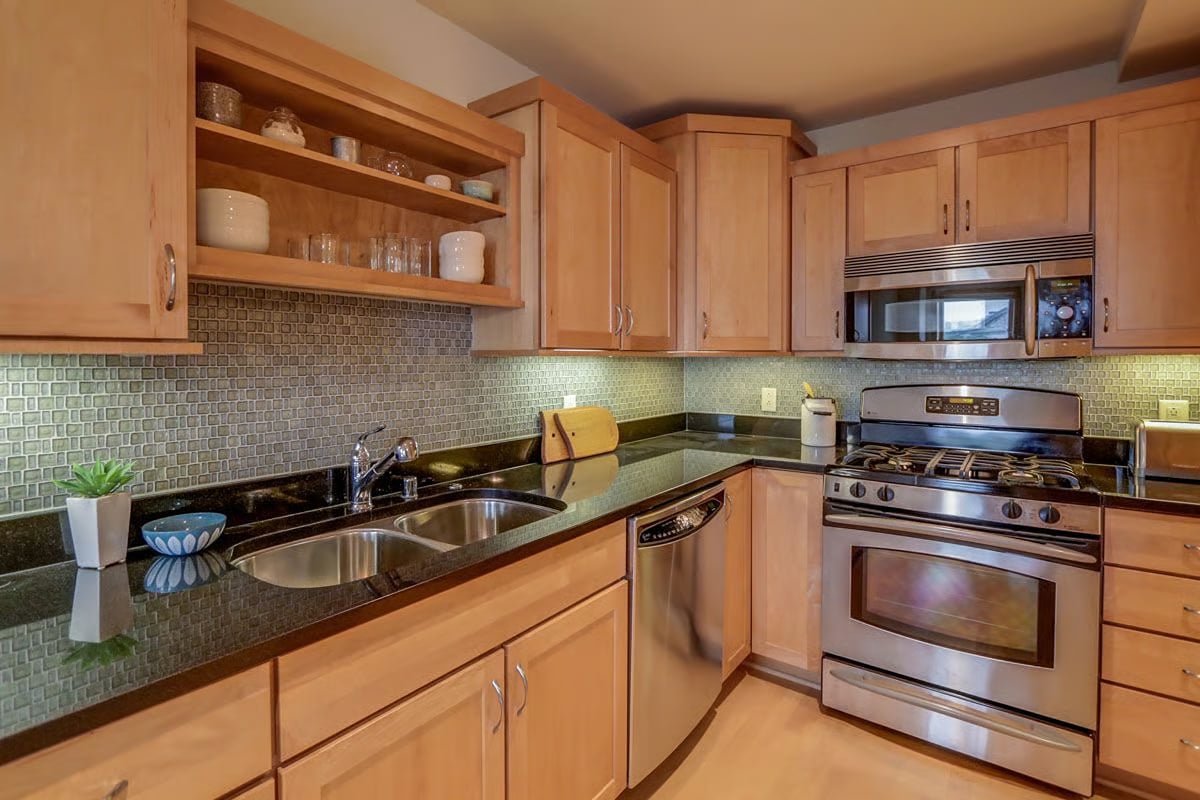 Kitchen with light wood cabinetry, black countertops, tiled backsplash, double sink, and stainless steel range.