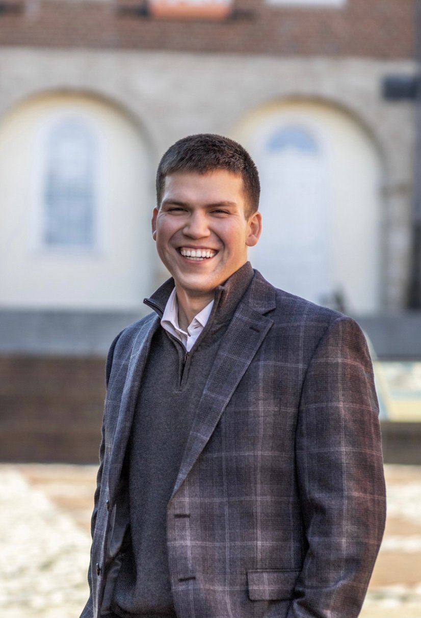 Theodosius Zotos smiling in a plaid blazer outdoors in front of a building with arched windows.
