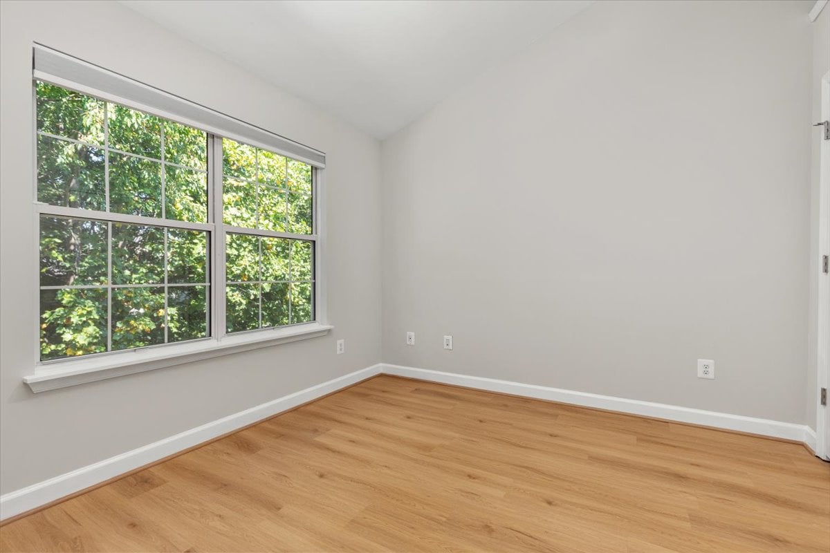 Empty room with light wood floors and large windows overlooking leafy trees.