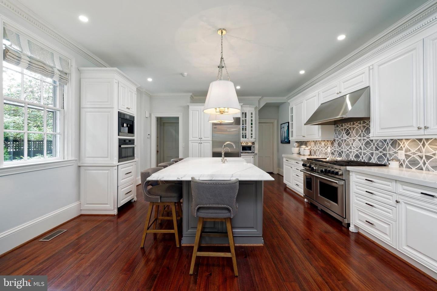 White kitchen with a large gray island, stainless steel range, patterned backsplash, and dark hardwood floors.