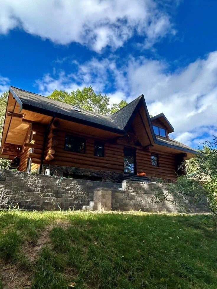 Log home with a steep gabled roof and stone retaining wall, viewed from below.