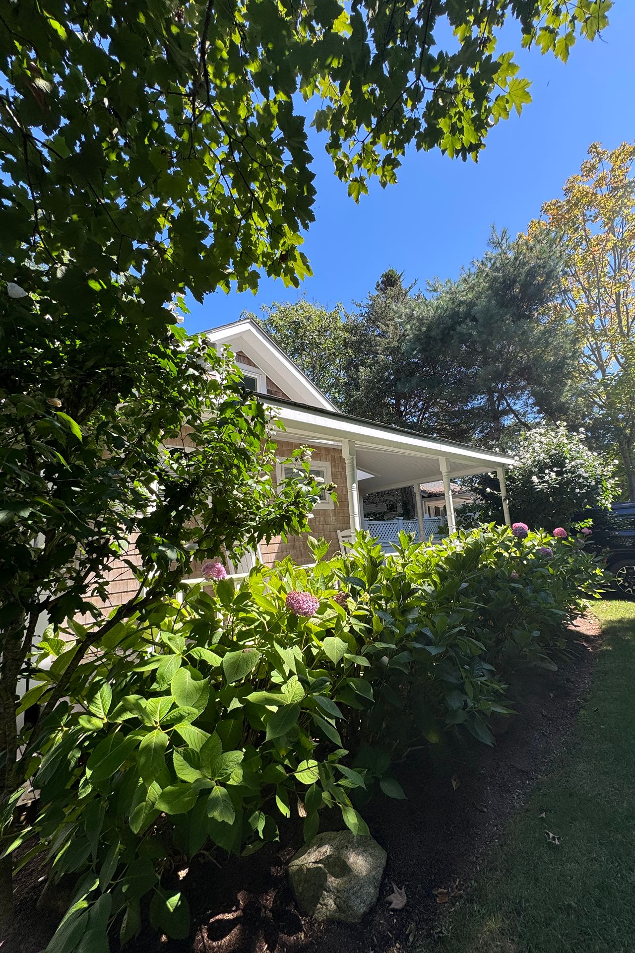 Shingled home with a covered porch, framed by leafy trees and pink hydrangeas.