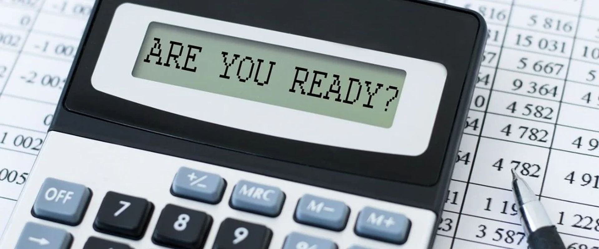Calculator displaying “ARE YOU READY?” on a printed spreadsheet beside a pen.