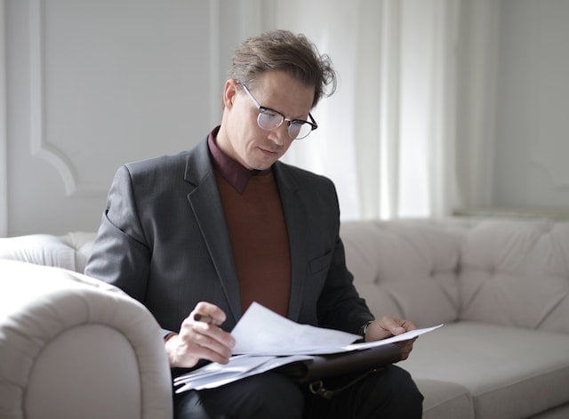A man in glasses and a suit sits on a beige sofa, focused on reading a lease agreement.