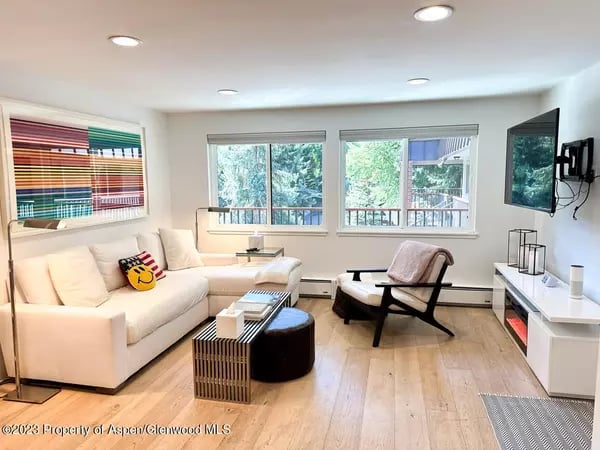 Living room with a white sectional, lounge chairs, light wood floors, and large windows overlooking trees.