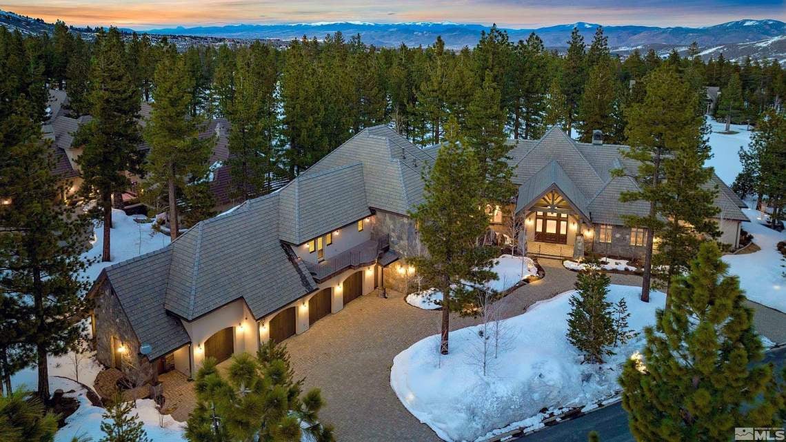 Aerial view of a large mountain home among snowy pines, with mountains in the distance at dusk.