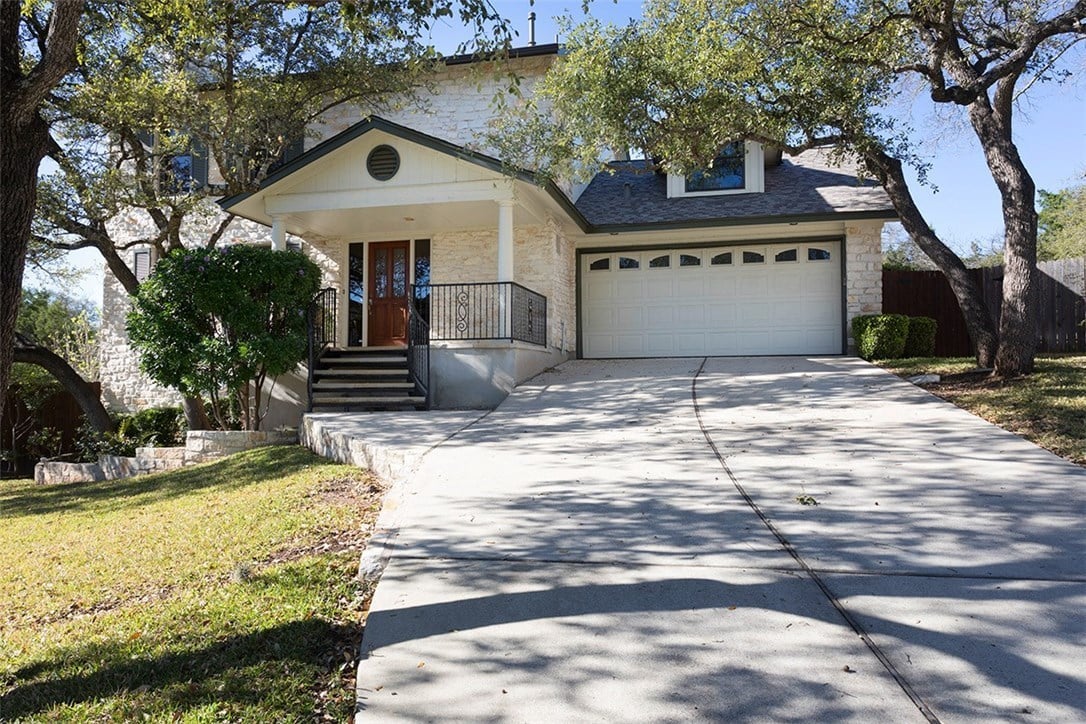 Light stone house with a covered entry, attached garage, and wide driveway beneath mature trees.