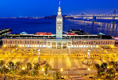 San Francisco Ferry Building illuminated at night with Bay Bridge in the background.