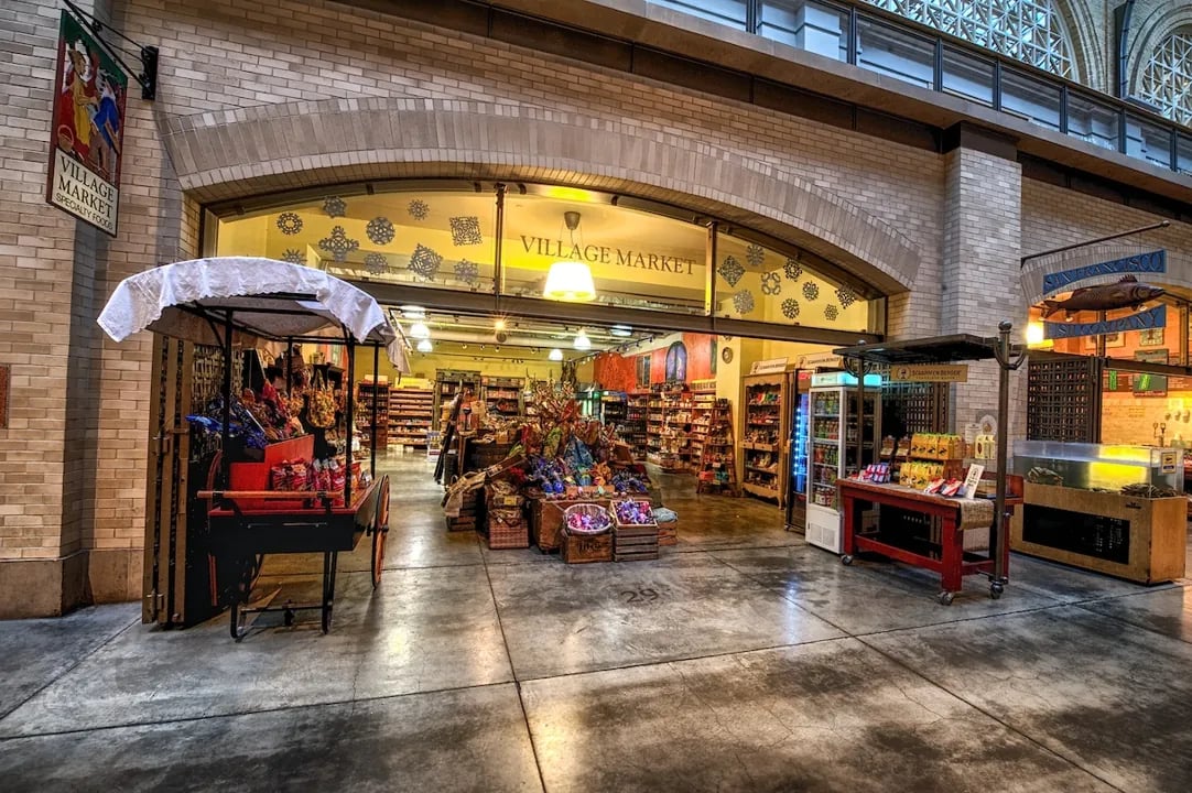 Village Market storefront in the Ferry Building with specialty foods signage and a red display cart.